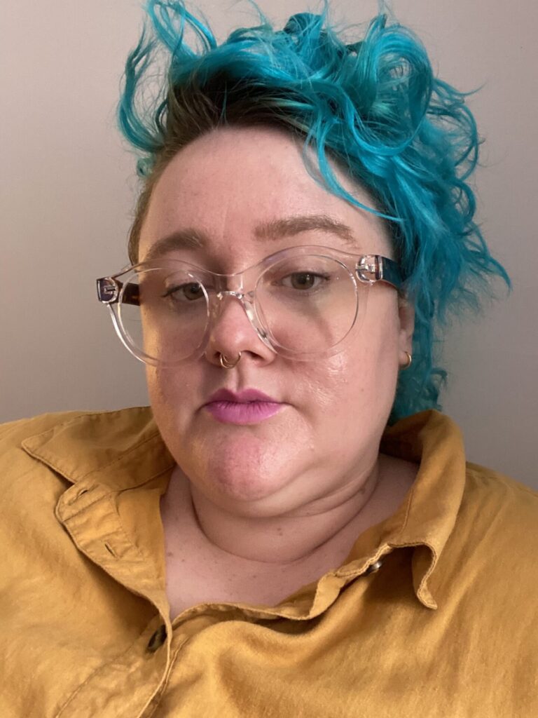 Close-up of JRE, a fat white femme, with blue hair, glasses, and in a button-down ochre shirt